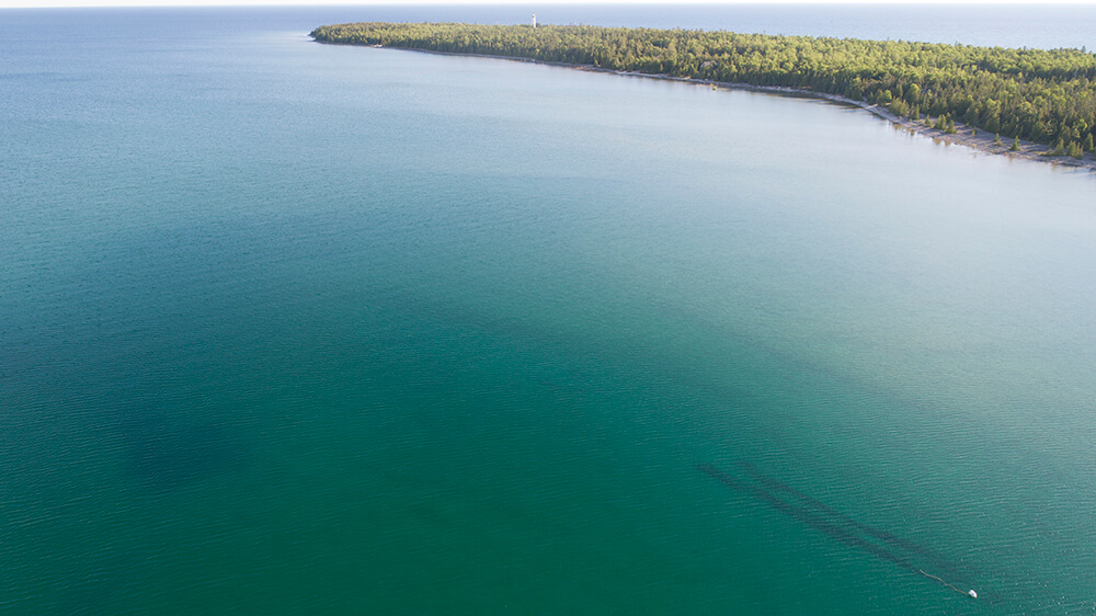 an aerial view shows a shipwreck below the surface and a lighthouse off in the distance