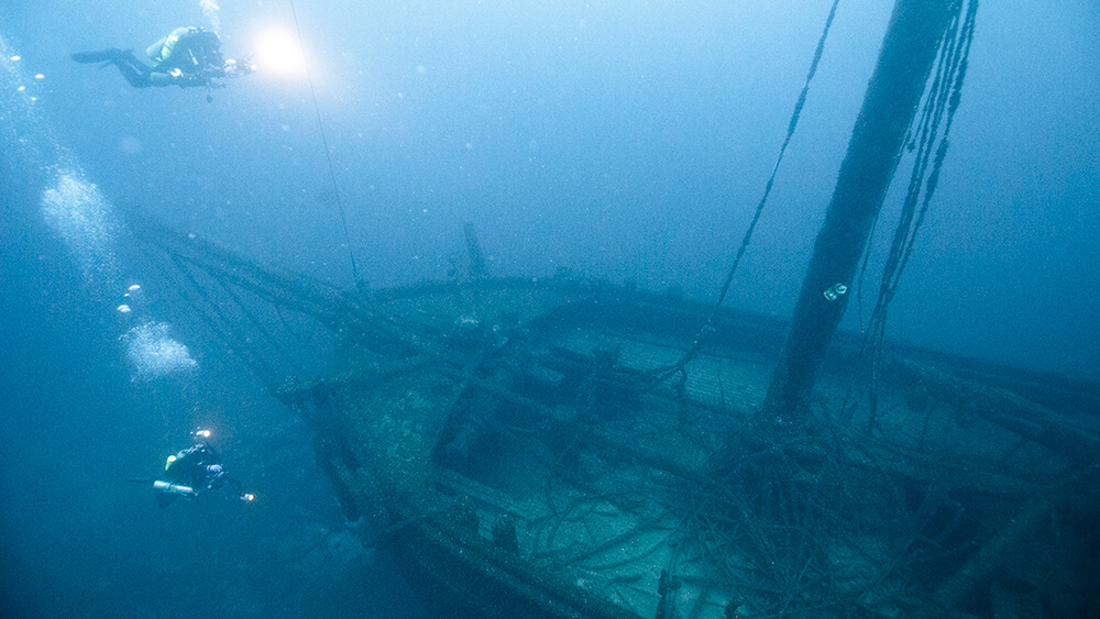 Two divers with bright lights swim above the deck of a wooden shipwreck