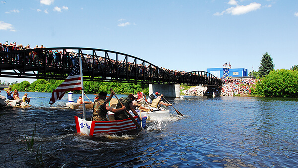 People paddle cardboard boats as a crowd watches from a bridge