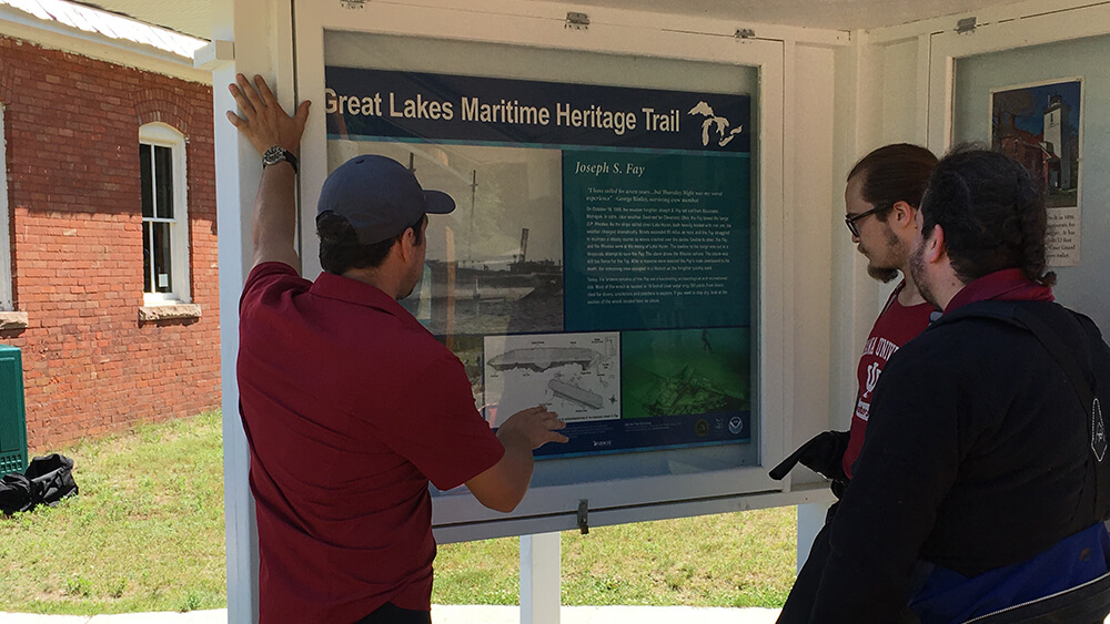 a man points at the diagram of a shipwreck on a trail sign
