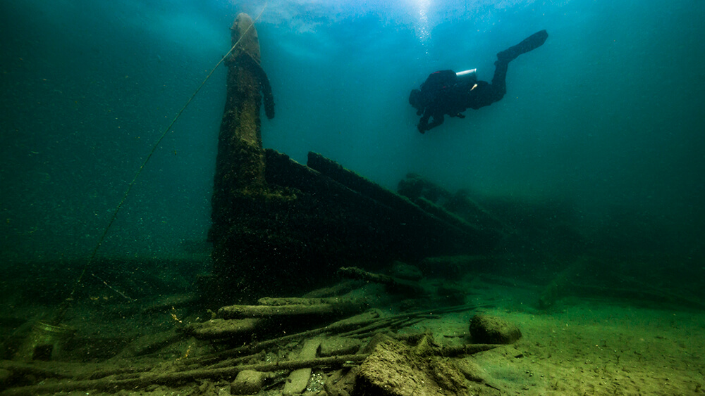 a diver swims above the bow of a shipwreck while light filters diwn from the surface
