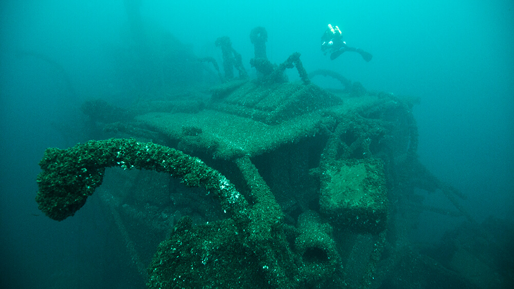 a diver swims above a shipwreck with twisted beams of metal prptruding form it
