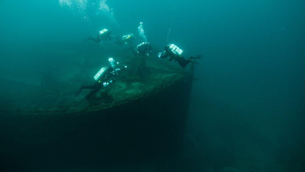 4 divers hover above the bow of a shipwreck