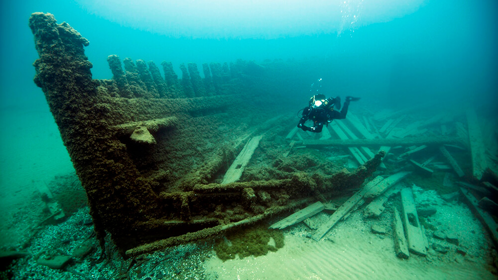 A diver swims near the bow of a shipwreck, most of the wood is missing from the near side